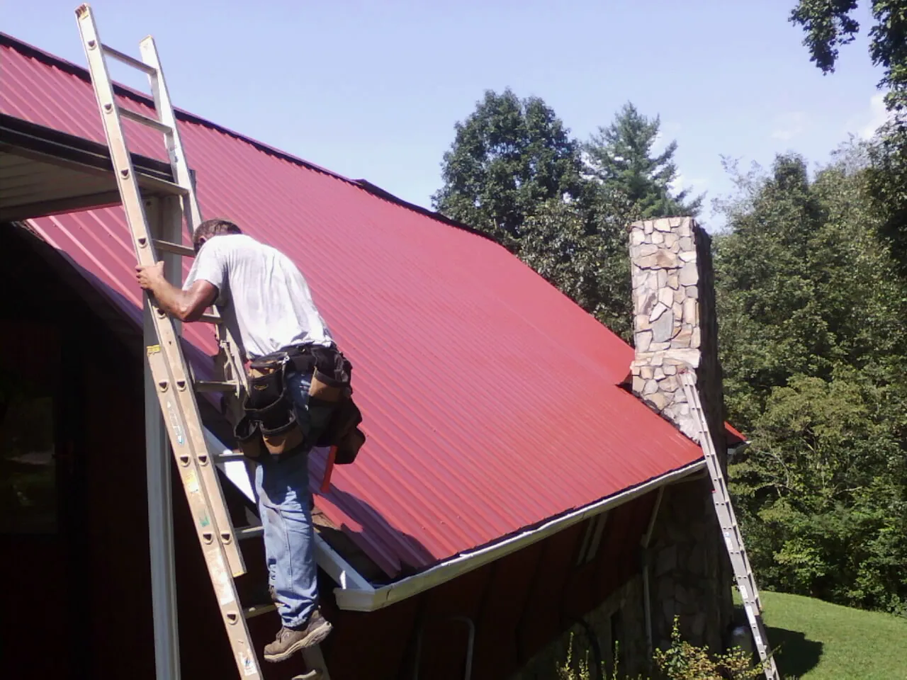 Roofer on a ladder beside a red metal roof and stone chimney — Soffit & Fascia Repair by Roofing Brothers in Fishtail